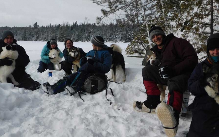 A group of people wearing heavy snow gear smile at the camera while they cuddle with sled dogs in a snowy landscape